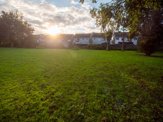 Wall Mural - a large open field with a houses estate in the background. the sun is setting and the sky is cloudy.