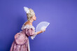 © deagreez - Elegant royal woman dressed in ornate baroque costume with fan posing against a vibrant purple background