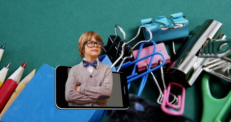 Poster - Image of smart caucasian schoolboy in smartphone moving over school stationery on desk