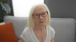 © Krakenimages.com - Elderly woman with albinism sitting in a cozy living room, wearing glasses and a white shirt, appears thoughtful in her home environment.