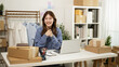 © saltdium - Portrait of Smiling young woman working at home on her online clothing shop. Surrounded by packages, shirts, and office tools