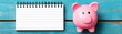 © Jirapong - Close-up of a pink piggy bank placed next to a blank spiral notebook on a bright turquoise wooden surface for saving money and financial planning concept