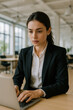 © JulieMeneghin - Femme en tailleur noir travaillant sur ordinateur portable dans un espace de coworking moderne, concentrée sur des tâches administratives ou de gestion d’entreprise