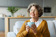 © Home-stock - Dreamy elderly woman enjoying good morning, drinking hot coffee while resting on comfortable couch in living room, looking away and smiling