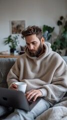 Poster - A young man in a cozy oversized hoodie focuses intently on his laptop while holding a warm cup of coffee. The room is bright and inviting, adorned with lush houseplants