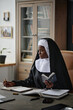 © AnnaStills - Vertical shot of young adult African American nun writing her thoughts in notepad while she sitting in brown leather office chair in office