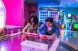 © Koldo_Studio - Two friends enjoying a competitive game of air hockey, surrounded by the vibrant lights and atmosphere of an entertainment venue