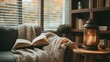 © sitifatimah - Book rests open on sofa covered in throw blanket, next to glowing lantern, pinecones, evoking warmth.