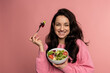 © Nataliya - Joyful cute young woman enjoying her favorite mouthwatering vegan dish in front of the camera on the pink background. Healthy food concept