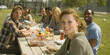 © ABF_MATEO - A diverse group of friends enjoys a picnic together outdoors on a sunny day, eating and smiling with a grassy field background.