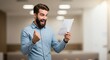 © ManuStockStudio - Bearded man in blue shirt holding paper with thumbs up and excited expression in blurred background
