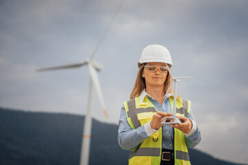 Poster - In a busy engineering setting, a female engineer in a hard hat inspects a miniature wind turbine model, with fullsized turbines in the background, showcasing advancements in renewable energy