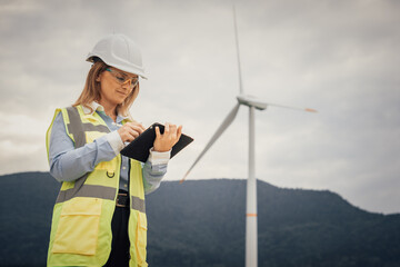 Poster - A female engineer in a safety helmet and yellow vest inspects data on a tablet at a wind energy site, surrounded by tall wind turbines representing advancements in renewable energy