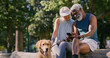 © Mitchell/peopleimages.com - Mature couple, dog and park with phone and web scroll with social media and pet outdoor. Morning, love and space of interracial relationship with animal, fitness break and tech with golden retriever