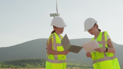 Fototapete - Two skilled female engineers in safety gear are reviewing documents for a wind turbine project, emphasizing teamwork and dedication to advancing renewable energy for a sustainable future