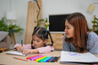 © kenchiro168 - Young girl drawing with colored markers while woman watches supportively at wooden table in cozy living room