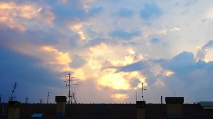 Poster - Bright yellow and orange hues dominate sky as sun sets, casting a vibrant glow over rooftops in the evening. Clouds reflect warm colors, creating serene atmosphere. Real time. Dramatic cumulus clouds