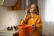 © Grustock - Worried woman collecting dripping water in orange bucket while calling plumber or insurance company to report ceiling water leak in kitchen