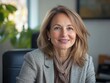 © Denis - A professional, confident woman with blonde hair, smiling at the camera and looking directly into it. She is wearing a business suit and a pearl necklace. The background features an office setting