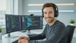 © f_bossa - A smiling young man with headphones sits at a desk in a modern office, working on two computer monitors displaying data visualizations.