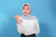 © Saddha Visual - Young Muslim woman wearing a grey hijab smiling brightly while glancing to the side and pointing with her thumb, showing a cheerful and friendly expression on blue background.
