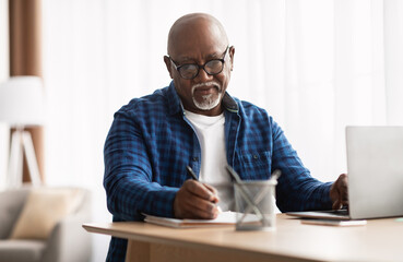 Entrepreneurship Career. Mature African American Man Taking Notes Working Sitting At Laptop Computer In Modern Office Indoors. Senior Businessman Writing Report At Workplace, Wearing Eyeglasses