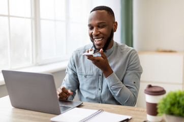 Poster - African Businessman With Smartphone Using Voice Assistant App Or Loudspeaker Talking Holding Phone Near Mouth Sitting At Workplace In Modern Office. IOT, Business Applications And Voice Search