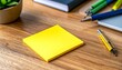 © Rendy Dastian - Yellow sticky note rests on wooden desk surrounded by pens and plant, creating vibrant workspace atmosphere.
