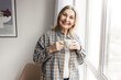 © shurkin_son - Senior Caucasian lady in casual clothes enjoying coffee in morning admiring cityscape through window, holding white mug in hands and smiling at camera. Lifestyle of retired people concept
