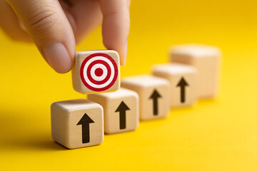 Poster - Hand placing wooden cube with target symbol atop cubes with upward arrows, symbolizing achieving goals and progress on yellow background