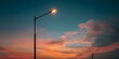© horizon - Single modern illuminated street lamp silhouetted against vibrant sunset sky featuring warm orange, pink, and twilight blue hues.
