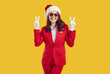 © Studio Romantic - Happy stylish woman Santa Claus in red hat, costume and sunglasses, gesturing victory or peace sign, expressing positive vibes, posing against yellow studio background. New Year concept