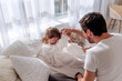 © farmuty - Father tickling his young daughter while she sits on a bed, both laughing joyfully in a brightly lit bedroom adorned with soft white curtains and cozy bedding
