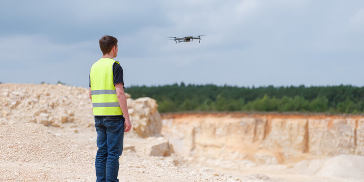 A person in a high-visibility vest operates a drone at an open quarry site with forest in the background. Concept: Technology and surveying in natural environments