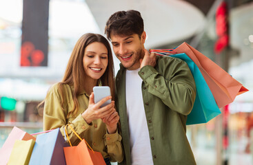 Poster - Happy Couple Shopping Using Mobile Phone Application Holding Colorful Shopper Bags And Buying Clothes In Modern Mall On Weekend. Customers Hold Smartphone Browsing Internet For Sales. Ecommerce
