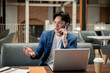 © bongkarn - Blue suit asian man talking on phone with explaining hand sitting with laptop in cafe or coffee shop