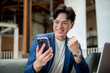 © bongkarn - Smiling asian man is making a fist while looking at phone sitting with laptop in cafe or coffee shop