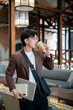 © bongkarn - Brown suit man with glasses drinking coffee and carrying laptop and notebook while standing in cafe.