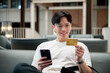 © bongkarn - Asian man wearing glasses in white tee holding phone and looking at credit card while sit on sofa.