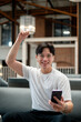 © bongkarn - Asian man wearing glasses in white tee is looking excited while holding his phone and raising a fist