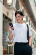 © bongkarn - Asian man with glasses in white tee walking hand in pocket and looking at phone in a mall or market
