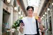 © bongkarn - Smiling asian man with glasses in white tee holding phone and showing credit card in shopping mall