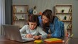 © kinomaster - Young child girl studying at home, small girl sits at desk and attends school class online on laptop, doing homework, mother helping and controlling process.