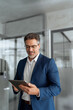 © Stock 4 You - Focused senior mature Indian or Latin entrepreneur businessman holding digital pc tablet standing in office. Smiling hispanic man in suit working using touchpad computer for business work. Vertical