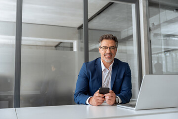 Portrait senior finance professional man working on cellphone telephone, pc laptop computer looking at camera. Mature ceo businessman in suit using phone app for business work in office. Copy space