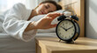 © abu - Woman Turning off Black Alarm Clock Lying on Bed in Bright Morning Light Wearing White Robe