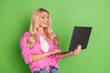 © deagreez - Young woman with blonde hair using a laptop against a vibrant green background, wearing a casual pink shirt