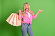 © deagreez - Excited Young Woman Holding Shopping Bags and Pointing Cheerfully Against a Vibrant Green Background Promoting
