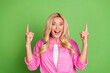 © deagreez - Joyful young woman with blonde hair smiling and pointing upwards while wearing a pink shirt against a green background