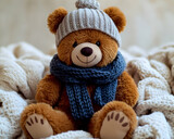 Plush brown teddy bear wearing a gray knit hat and dark blue scarf sitting on a fluffy white knit blanket